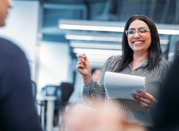 Woman speaking to a group with a smile and a pen in her hand 