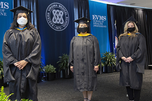 Three women graduates in gowns stand in front of the EVMS seal. All are wearing masks.