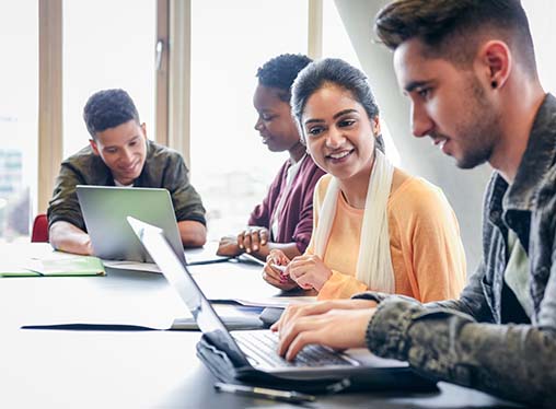 Three young men and one woman sit at a table using laptop computers.