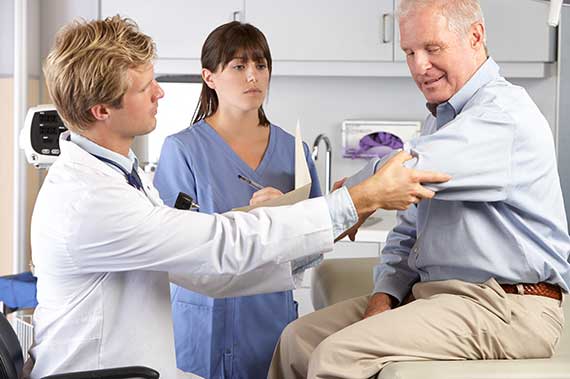 A physician examines a patient's arm while a student observes in the exam room.